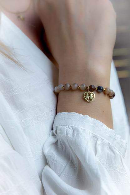 Ręka w białej bluzce z haftem, nosząca bransoletkę Serce Rodziny z agatów w odcieniach beżu i brązu z złotym charms.