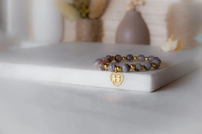 Bransoletka Serce Rodziny Agat Złoto z dominującymi szaro-białymi agatami przeplatanymi złotymi koralikami, ozdobiona złotą zawieszką w kształcie serca z sylwetkami dzieci. Produkt ułożony na białej powierzchni, w tle subtelne dekoracje.