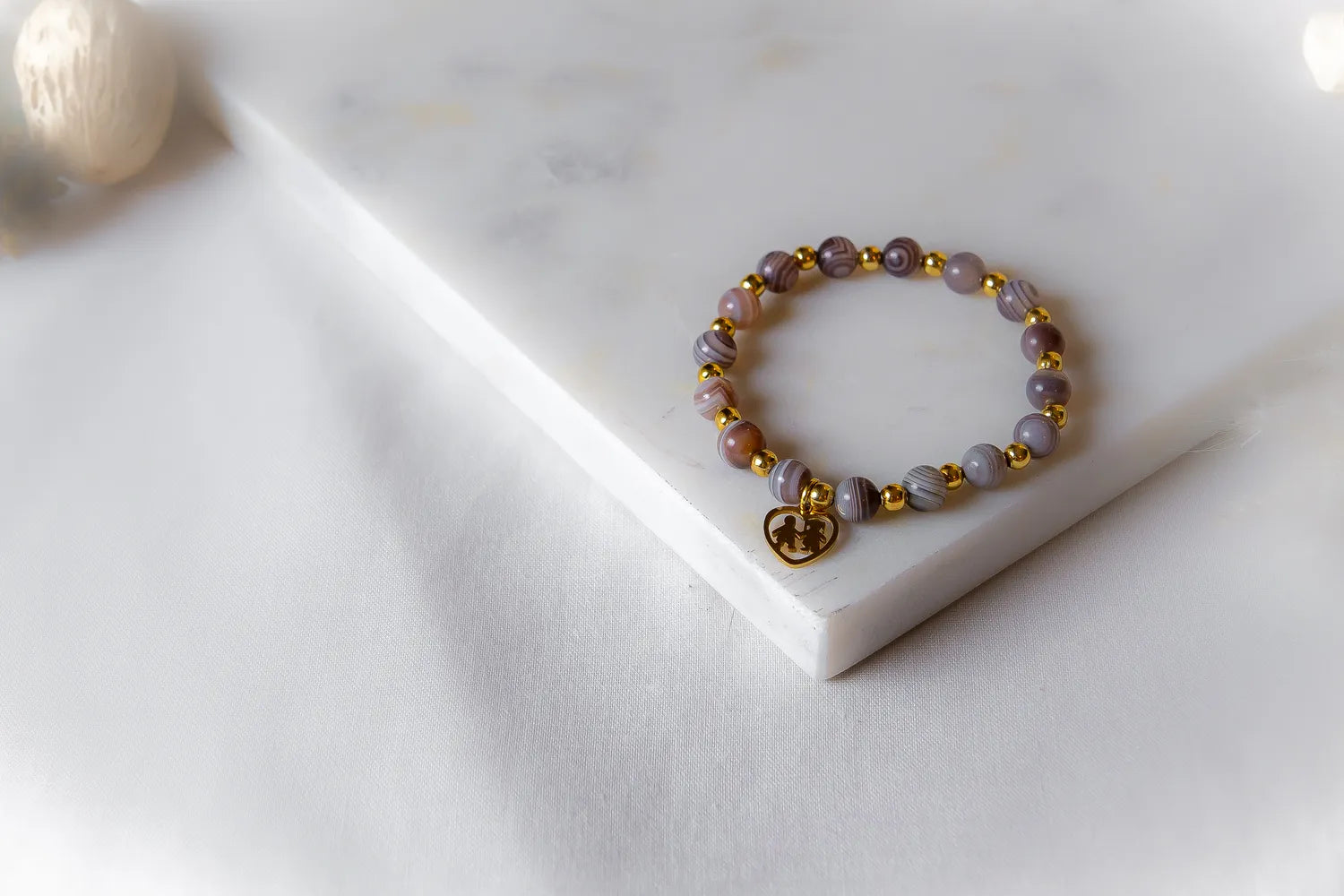 Serce Rodziny Bransoletka Agat Złoto z błyszczącym złotym zawieszką w kształcie serca, na tle białej tkaniny i marmuru.