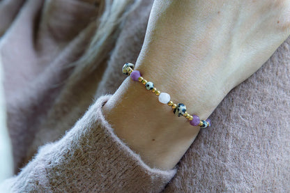 SŁODKI IMPULS - BRANSOLETKA Z JASPISU DALMATYŃSKIEGO I JADEITU. Bransoletka z kolorowymi kamieniami, w tym jaspisem dalmatyńskim i jadeitem, na tle delikatnej, beżowej bluzy. Ułożenie na nadgarstku.