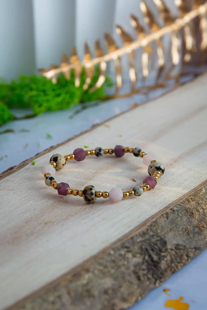 SŁODKI IMPULS - BRANSOLETKA Z JASPISU DALMATYŃSKIEGO I JADEITU. Bransoletka z kolorowymi koralikami jaspisu dalmatyńskiego i jadeitu, ułożona na drewnianym tle z zielonymi dodatkami w tle.