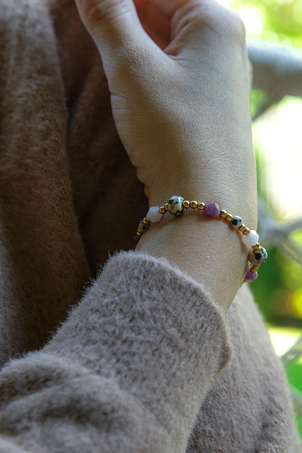 SŁODKI IMPULS - BRANSOLETKA Z JASPISU DALMATYŃSKIEGO I JADEITU na nadgarstku, złożona z kolorowych kamieni i złotych elementów, na tle miękkiego swetra.