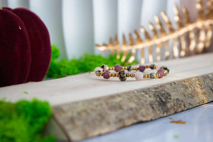 SŁODKI IMPULS - BRANSOLETKA Z JASPISU DALMATYŃSKIEGO I JADEITU, prezentująca się na drewnianym tle z zielonym mchem i czerwonym aksamitnym podkładem. Biżuteria składa się z kolorowych kamieni jaspisu dalmatyńskiego i jadeitu, ułożonych w elegancki wzór.