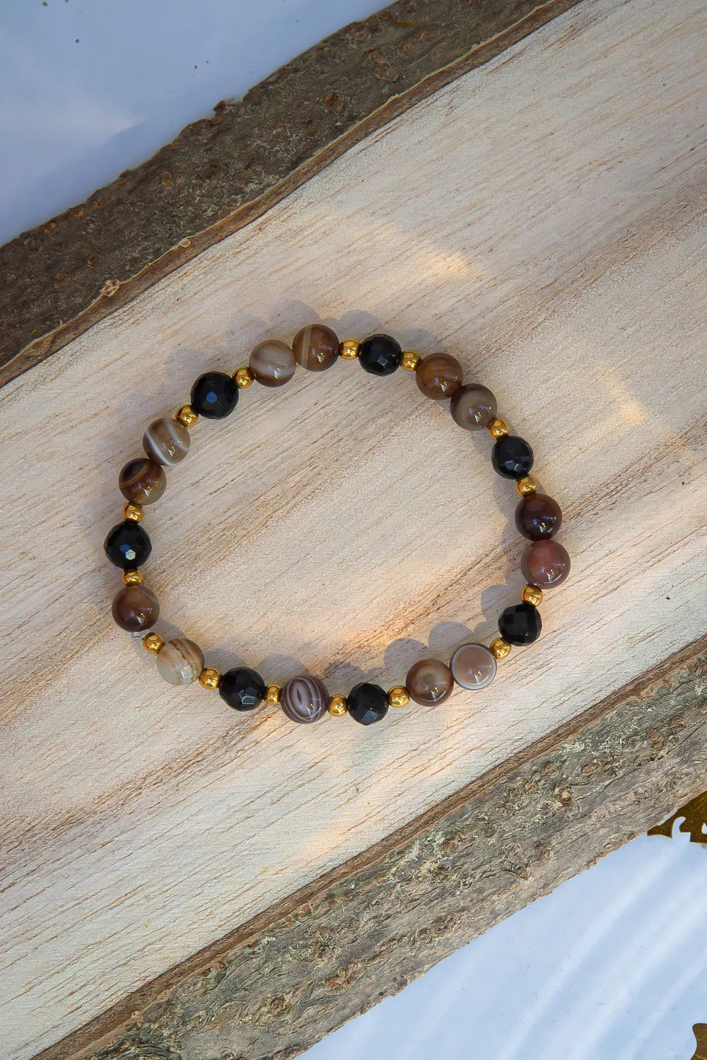 STRAŻNIK ENERGII - BRANSOLETKA Z TURMALINU I AGATU BOTSWANA. Bransoletka z koralikami turmalinu i agatu Botswana ułożona na drewnianym tle.