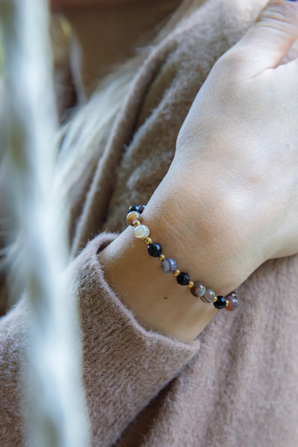 Bransoletka 'STRAŻNIK ENERGII - BRANSOLETKA Z TURMALINU I AGATU BOTSWANA' z widocznymi kolistymi kamieniami turmalinu i agatu botswana, noszona na nadgarstku kobiety w beżowym swetrze, tło z delikatnymi roślinnymi elementami.