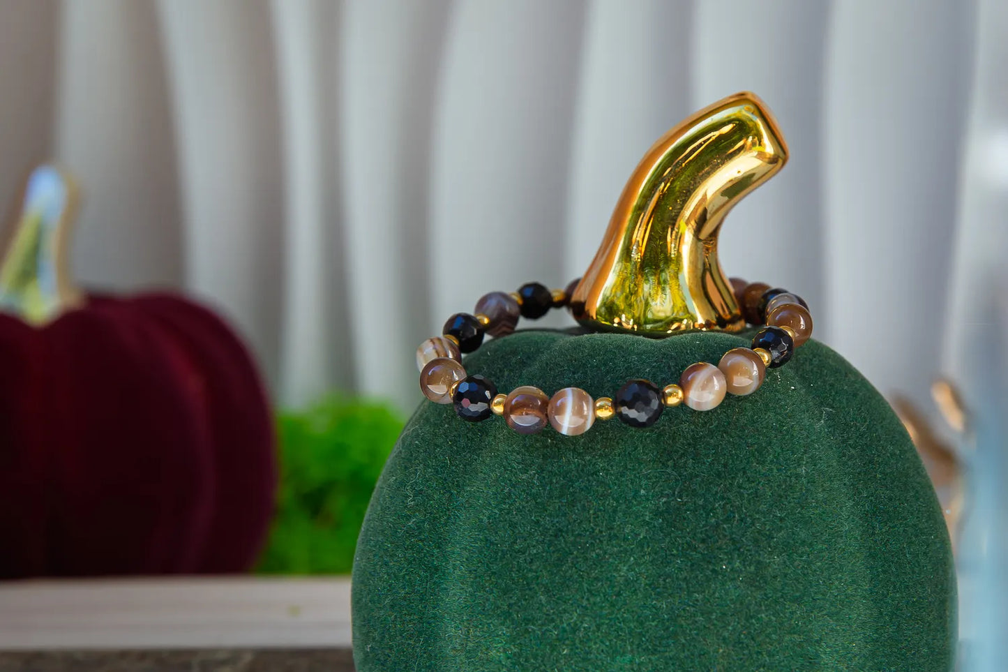 Bransoletka 'STRAŻNIK ENERGII - BRANSOLETKA Z TURMALINU I AGATU BOTSWANA' z koralikami turmalinu i agatu botswana, umieszczona na zielonym podstawce w kształcie dyni, tło z dyni i zasłoną.