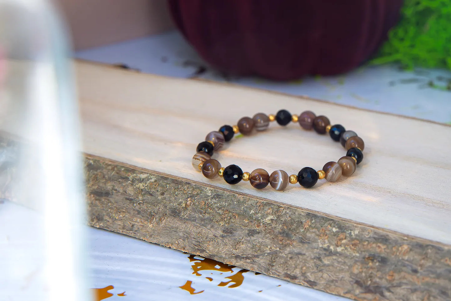 STRAŻNIK ENERGII - BRANSOLETKA Z TURMALINU I AGATU BOTSWANA, złożona z kolorowych koralików turmalinu i agatu, leżąca na drewnianym bloku z naturalnym tłem.