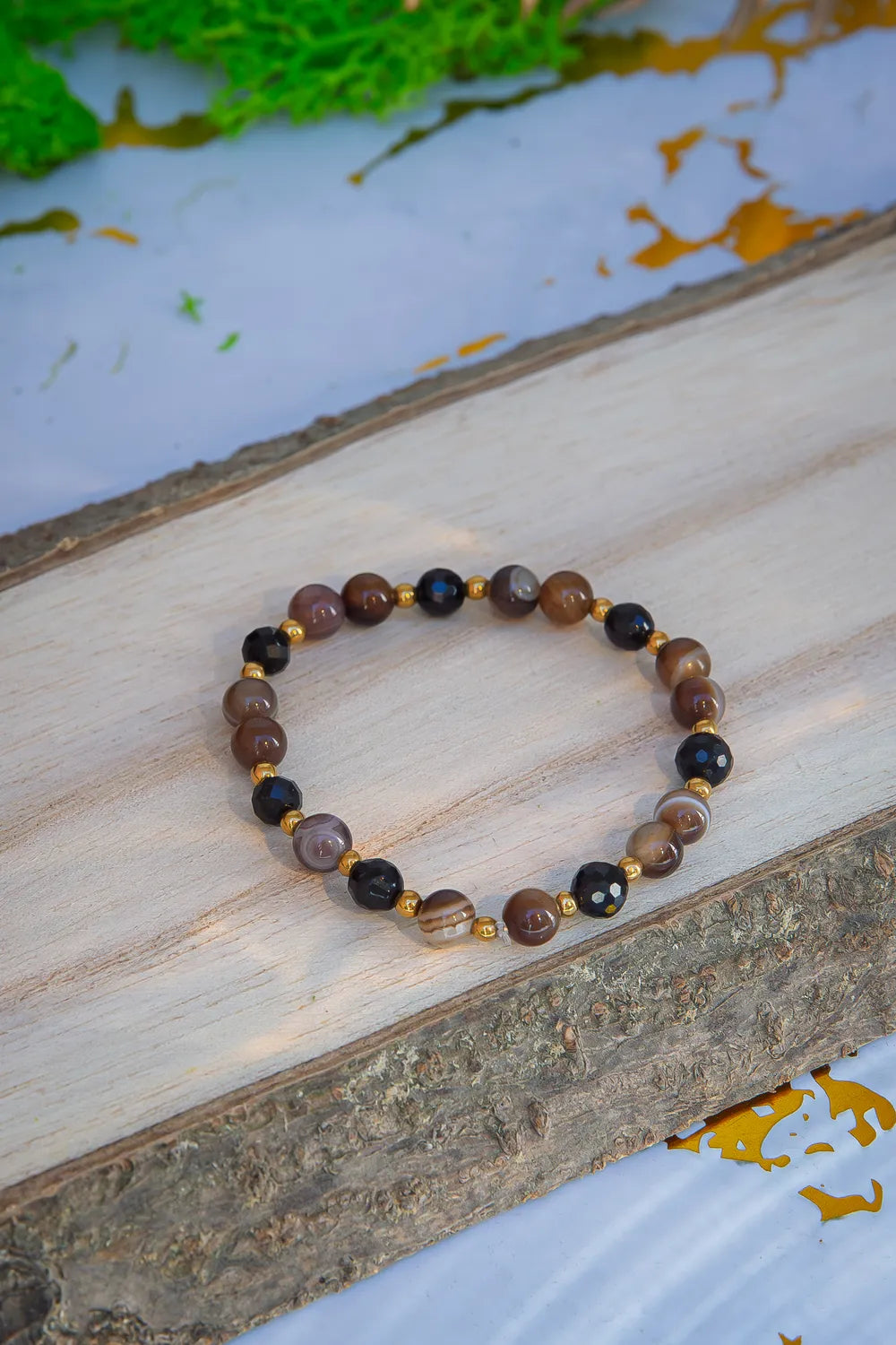 Bransoletka 'STRAŻNIK ENERGII - BRANSOLETKA Z TURMALINU I AGATU BOTSWANA' z kolorowymi kamieniami turmalinu i agatu botswana, ułożona na drewnianym tle z zielonymi liśćmi w tle.