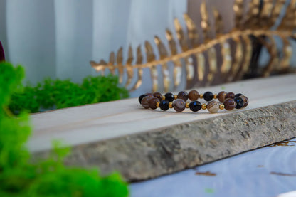 STRAŻNIK ENERGII - BRANSOLETKA Z TURMALINU I AGATU BOTSWANA, bransoletka z koralikami turmalinu i agatu Botswana, ułożona na drewnianym tle z zielonymi roślinami i złotym liściem w tle.