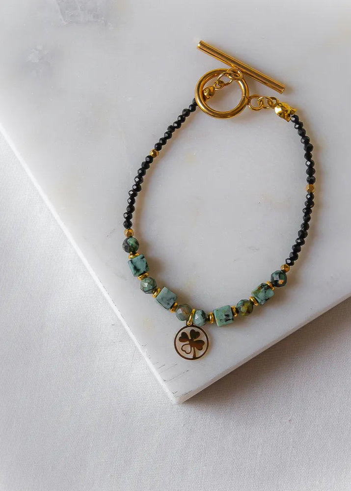 Szczęśliwa Koniczynka Bransoletka Z Turmalinu Czarnego I Turkusu Afrykańskiego: złota bransoletka z turkusowymi, sześciennymi kamieniami i czarnymi, drobnymi koralikami, zwieńczona zawieszką w kształcie czterolistnej koniczynki.
