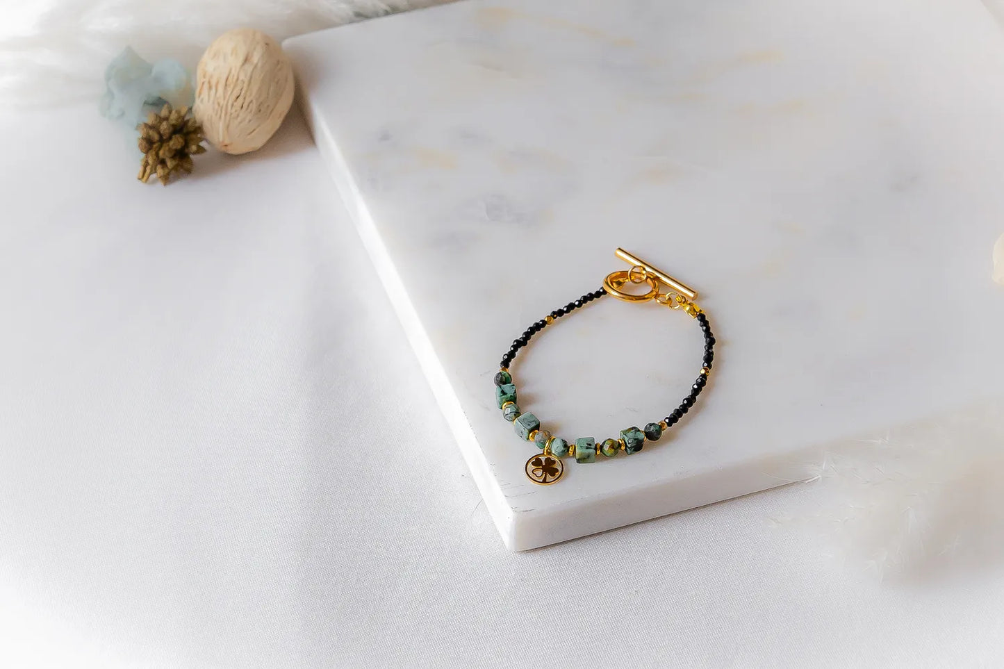 Szczęśliwa Koniczynka Bransoletka z Turmalinu Czarnego I Turkus Afrykańskiego Marmur: złota klamra, czarne koraliki, turkusowe, zielono-niebieskie kostki, zawieszka koniczynka, kamienie na białym tle marmuru.