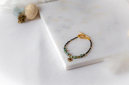 Szczęśliwa Koniczynka Bransoletka z Turmalinu Czarnego I Turkus Afrykańskiego Marmur: złota klamra, czarne koraliki, turkusowe, zielono-niebieskie kostki, zawieszka koniczynka, kamienie na białym tle marmuru.