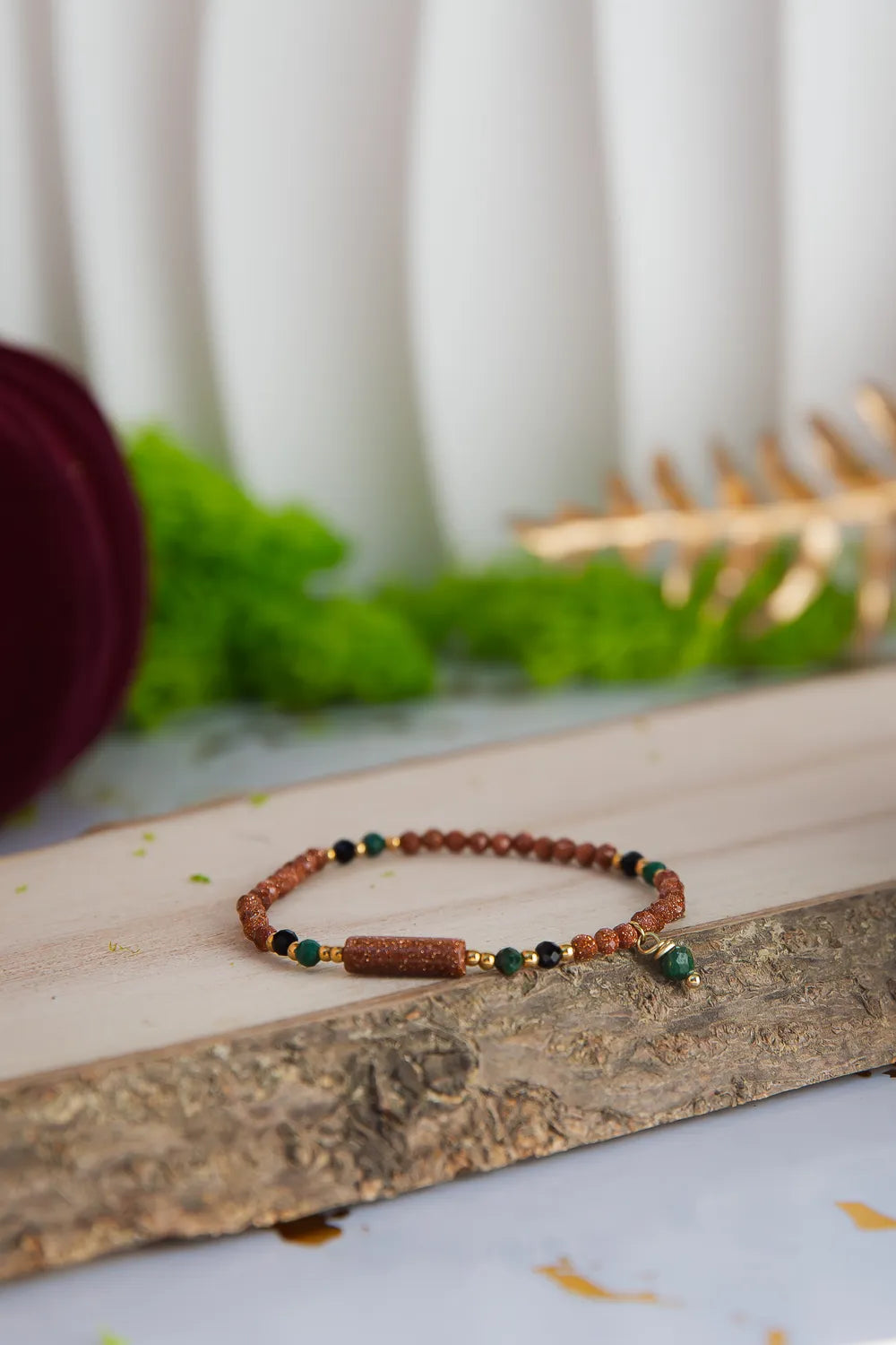 SZEPT LASU - BRANSOLETKA Z PIASKU PUSTYNI, AGATU I TURMALINU. Bransoletka z kolorowymi kamieniami, w tym piaskiem pustyni, agatem i turmalinem, leżąca na drewnianym pniu z zielonym tłem i dekoracją.