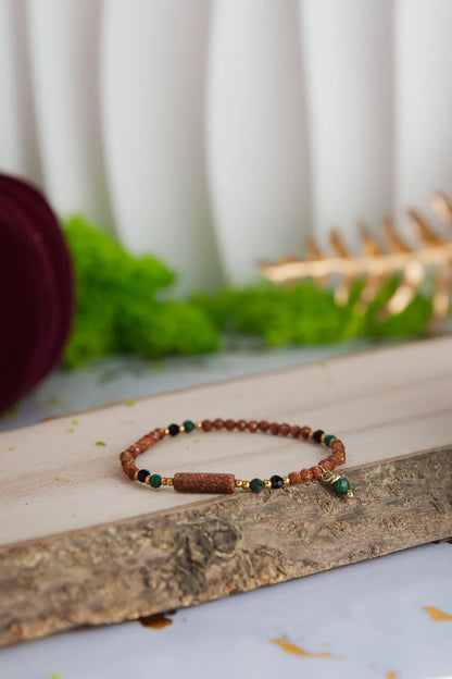 SZEPT LASU - BRANSOLETKA Z PIASKU PUSTYNI, AGATU I TURMALINU. Bransoletka z kolorowymi kamieniami, w tym piaskiem pustyni, agatem i turmalinem, leżąca na drewnianym pniu z zielonym tłem i dekoracją.
