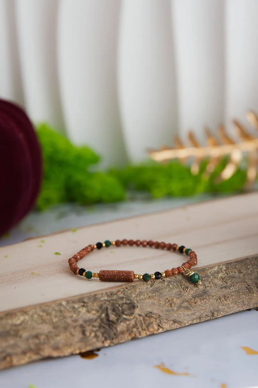 SZEPT LASU - BRANSOLETKA Z PIASKU PUSTYNI, AGATU I TURMALINU. Bransoletka z kolorowymi kamieniami, w tym piaskiem pustyni, agatem i turmalinem, leżąca na drewnianym pniu z zielonym tłem i dekoracją.