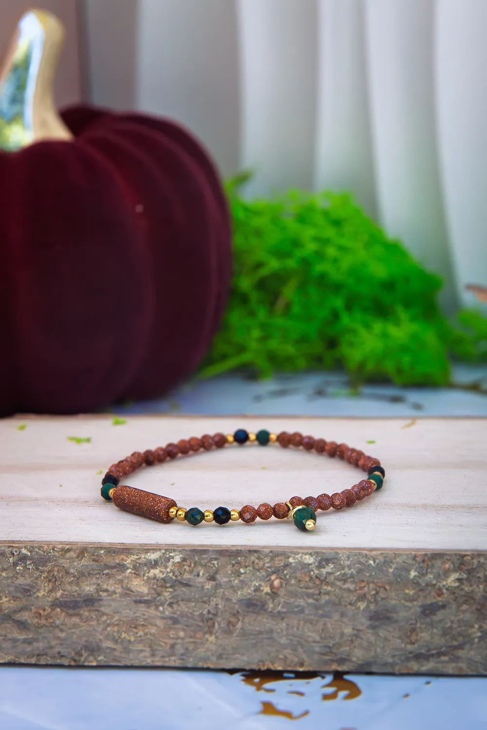 SZEPT LASU - BRANSOLETKA Z PIASKU PUSTYNI, AGATU I TURMALINU. Bransoletka z kolorowymi koralikami z piasku pustyni, agatu i turmalinu leży na drewnianym bloku. W tle widoczna czerwona dynia i zielone mchy.