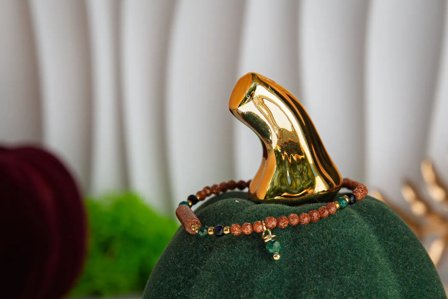 SZEPT LASU - BRANSOLETKA Z PIASKU PUSTYNI, AGATU I TURMALINU. Bransoletka z kolorowymi koralikami i zawieszką, umieszczona na zielonym aksamitnym podstawce z złotym dekoracyjnym elementem w tle.