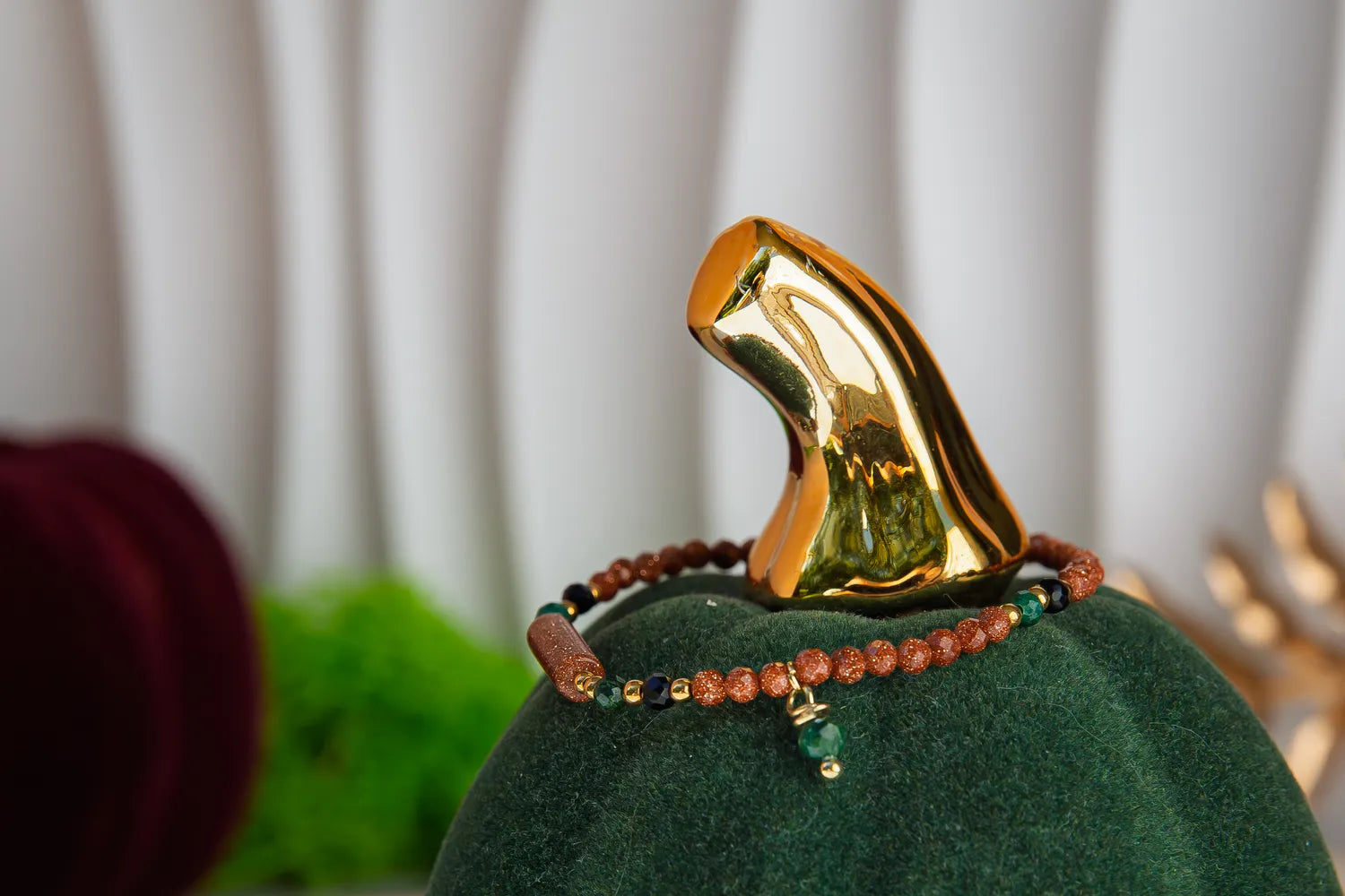 SZEPT LASU - BRANSOLETKA Z PIASKU PUSTYNI, AGATU I TURMALINU. Bransoletka z kolorowymi koralikami i zawieszką, umieszczona na zielonym aksamitnym podstawce z złotym dekoracyjnym elementem w tle.