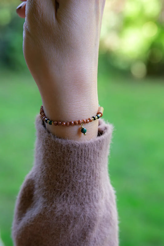 SZEPT LASU - BRANSOLETKA Z PIASKU PUSTYNI, AGATU I TURMALINU na nadgarstku, z widocznymi koralikami z piasku pustyni, agatu i turmalinu, na tle zielonej trawy.