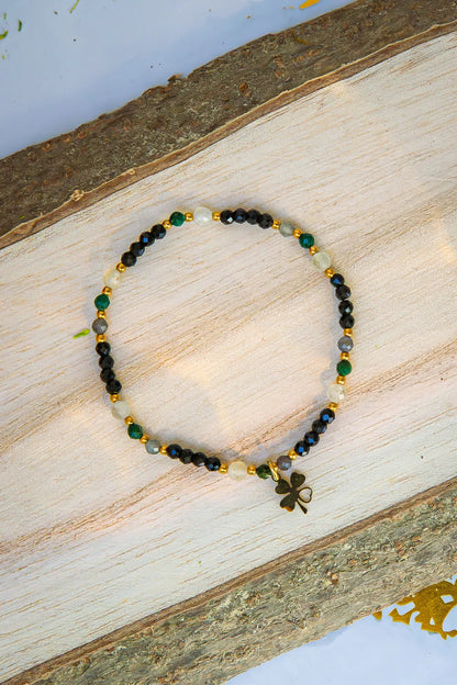 SZEPT SZMARAGDOWEJ WYSPY - BRANSOLETKA Z LEPIDOLITU, CYTRYNU I LABRADORYTU. Biżuteria z kamieni naturalnych i złotą koniczynką ułożona w koło na drewnianym tle.