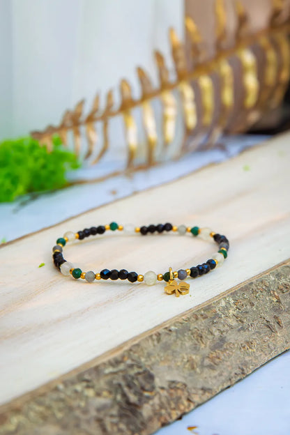 SZEPT SZMARAGDOWEJ WYSPY - BRANSOLETKA Z LEPIDOLITU, CYTRYNU I LABRADORYTU. Kolorowa bransoletka z kamieni ze złotym symbolem szczęścia na drewnianej powierzchni.