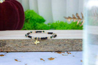 SZEPT SZMARAGDOWEJ WYSPY - BRANSOLETKA Z LEPIDOLITU, CYTRYNU I LABRADORYTU. Bransoletka szczęścia z koniczynką prezentowana na drewnianej belce w otoczeniu zieleni.