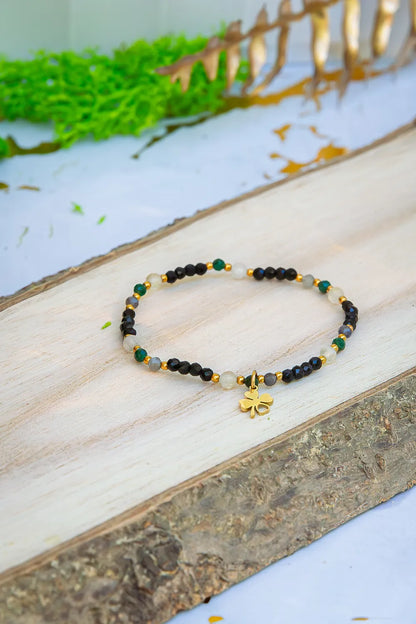 SZEPT SZMARAGDOWEJ WYSPY - BRANSOLETKA Z LEPIDOLITU, CYTRYNU I LABRADORYTU. Kompozycja z bransoletką z kamieni naturalnych i złotą koniczyną na drewnie z roślinami w tle.