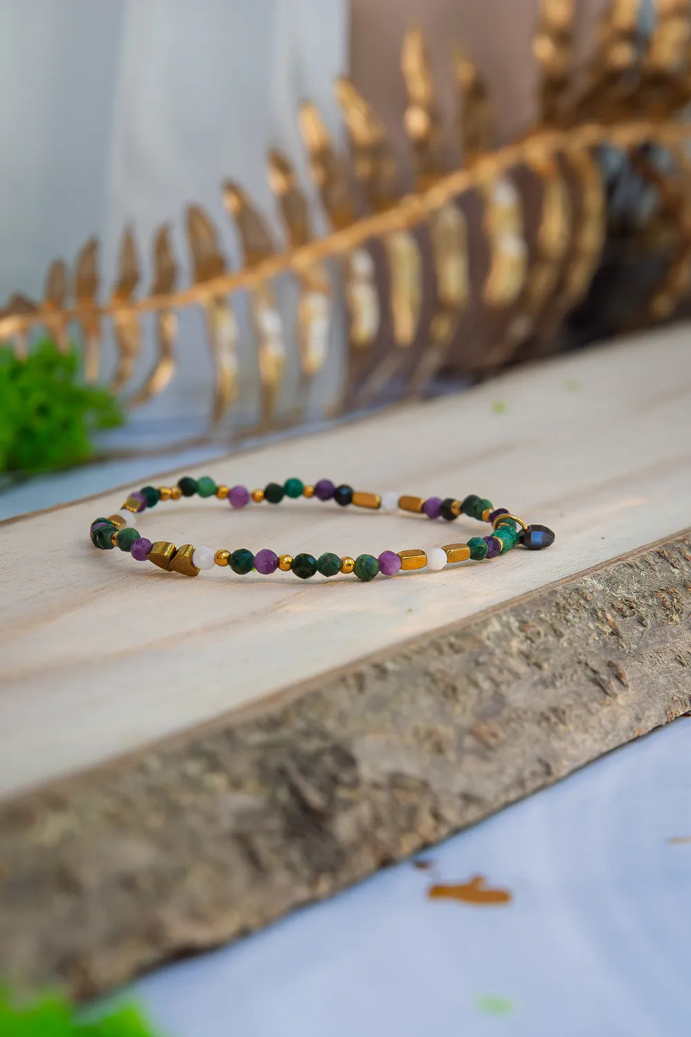 TAJEMNY OGRÓD - BRANSOLETKA Z LEPIDOLITU I CHALCEDONU leży na drewnianym pomoście z widocznymi kolorowymi koralikami z lepidolitu i chalcedonu, na tle złotej liściny i zielonych liści.