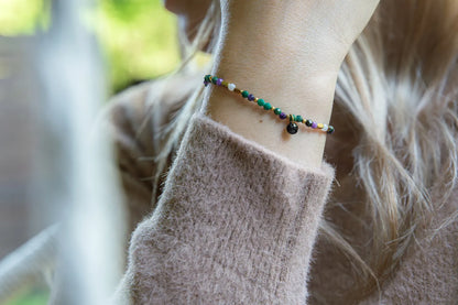 TAJEMNY OGRÓD - BRANSOLETKA Z LEPIDOLITU I CHALCEDONU na nadgarstku, złożona z kolorowych koralików i czarnego elementu, na tle delikatnej bluzki i rozmytego tła.