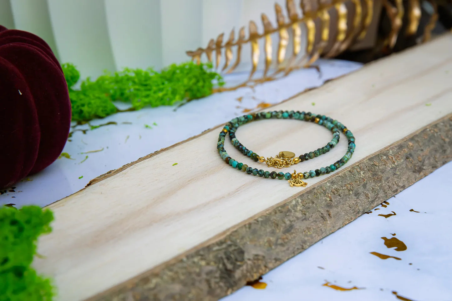 TARCZA ŻÓŁWIA - NASZYJNIK Z TURKUSU AFRYKAŃSKIEGO, złożony z małych, kolorowych kamieni turkusowych, leżący na drewnianym pomoście z zielonym mchem i złotymi liśćmi w tle.