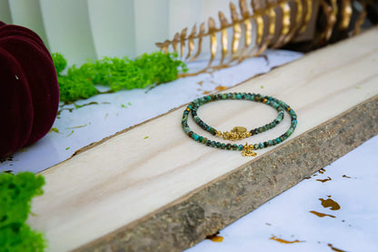 TARCZA ŻÓŁWIA - NASZYJNIK Z TURKUSU AFRYKAŃSKIEGO, złożony z małych, kolorowych kamieni turkusowych, leżący na drewnianym pomoście z zielonym mchem i złotymi liśćmi w tle.