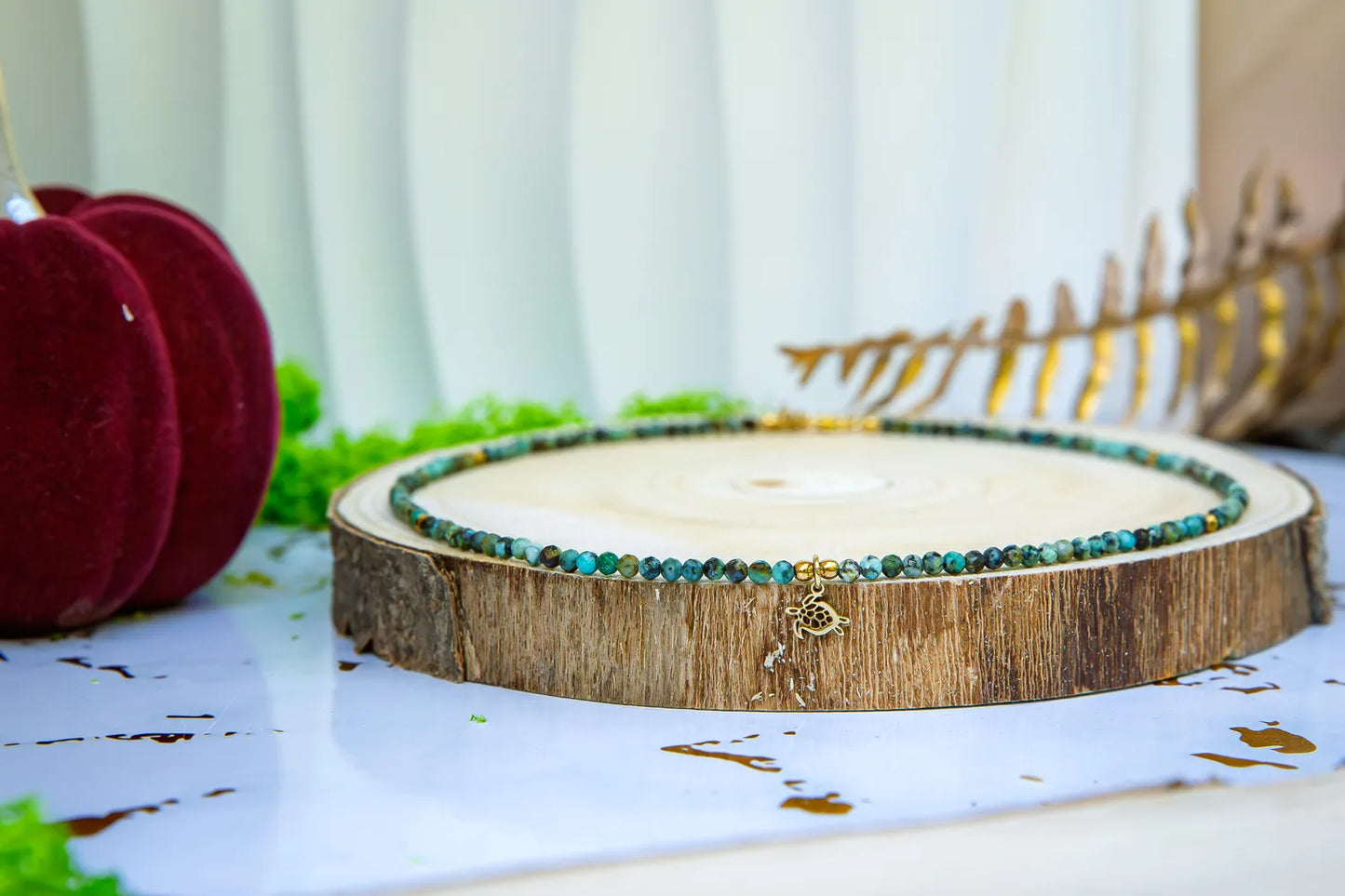 TARCZA ŻÓŁWIA - NASZYJNIK Z TURKUSU AFRYKAŃSKIEGO, elegancki naszyjnik z kolorowymi kamieniami turkusowymi, ułożony na drewnianym podstawce, tło z zielonymi liśćmi i czerwonymi ozdobami.