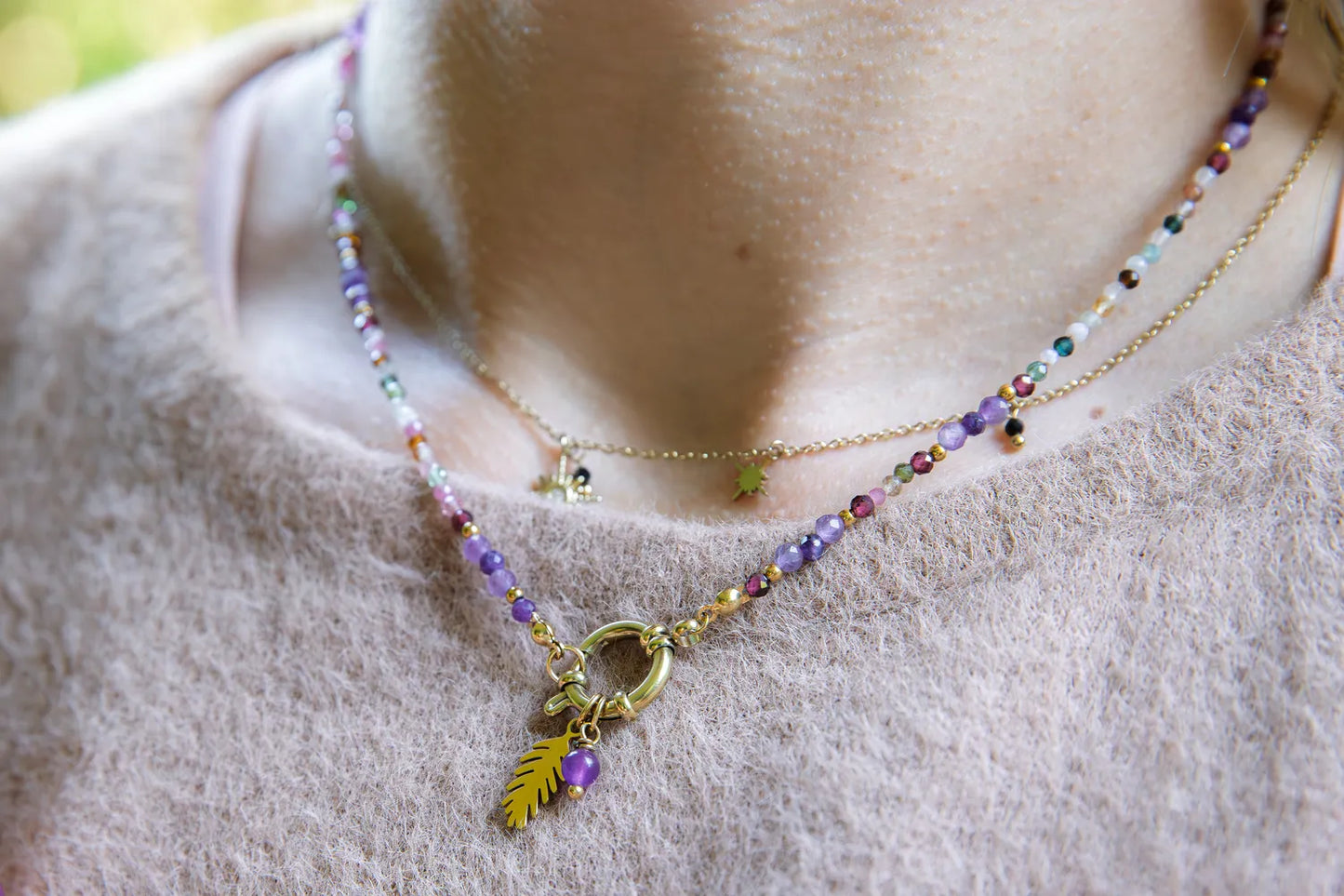 TĘCZOWA AURA - NASZYJNIK Z KOLOROWEGO TURMALINU I GRANATU. Złoty naszyjnik z kolorowymi kamieniami turmalinu i granatu, ozdobiony złotym liściem. Tło z delikatnej, beżowej tkaniny.