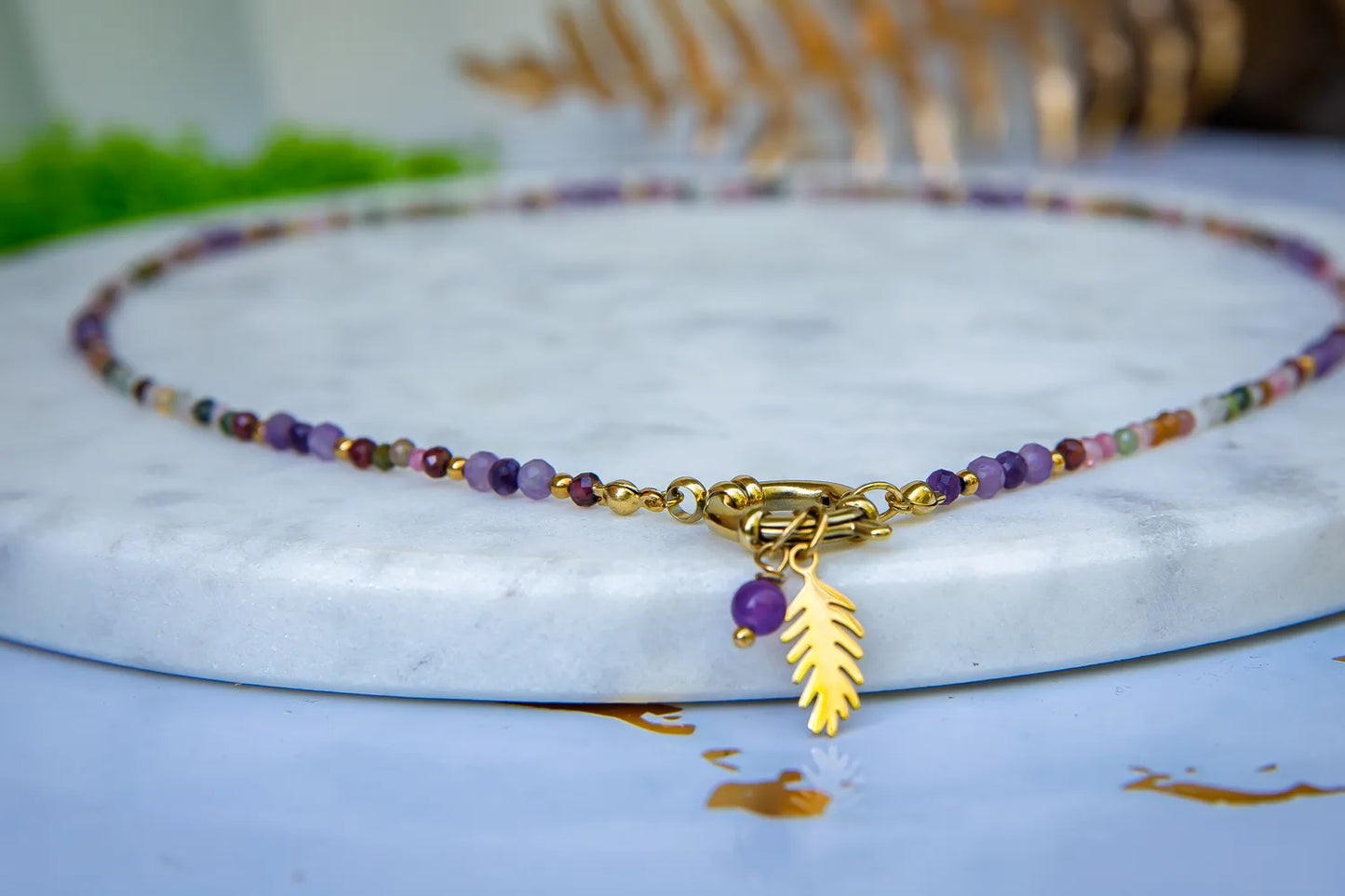 TĘCZOWA AURA - NASZYJNIK Z KOLOROWEGO TURMALINU I GRANATU. Naszyjnik z kolorowymi koralikami turmalinu i granatu, ozdobiony złotym zawieszką w kształcie liścia, leżący na marmurowym tle.