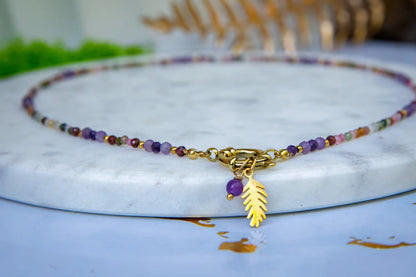 TĘCZOWA AURA - NASZYJNIK Z KOLOROWEGO TURMALINU I GRANATU. Naszyjnik z kolorowymi koralikami turmalinu i granatu, ozdobiony złotym zawieszką w kształcie liścia, leżący na marmurowym tle.