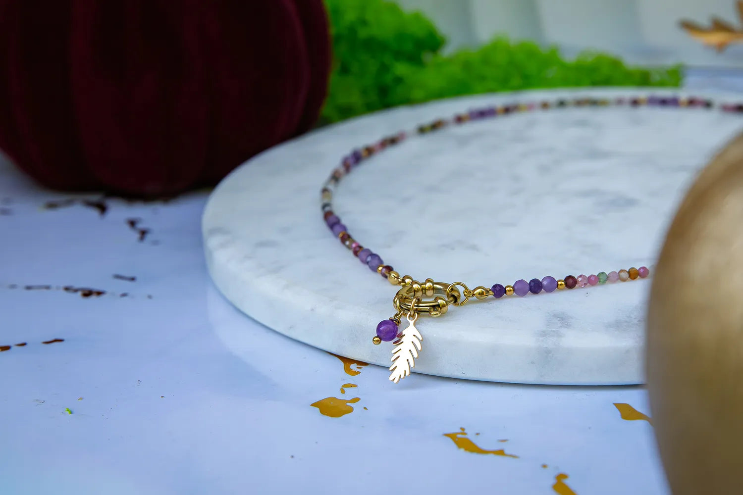 TĘCZOWA AURA - NASZYJNIK Z KOLOROWEGO TURMALINU I GRANATU. Naszyjnik z kolorowymi koralikami turmalinu i granatu, ozdobiony złotym liściem, na białym marmurowym tle.