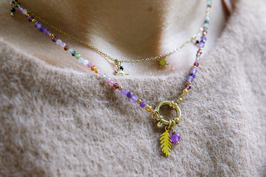 TĘCZOWA AURA - NASZYJNIK Z KOLOROWEGO TURMALINU I GRANATU. Naszyjnik z kolorowymi kamieniami turmalinu i granatu, ozdobiony złotym liściem i pierścieniem, na tle beżowego swetra.