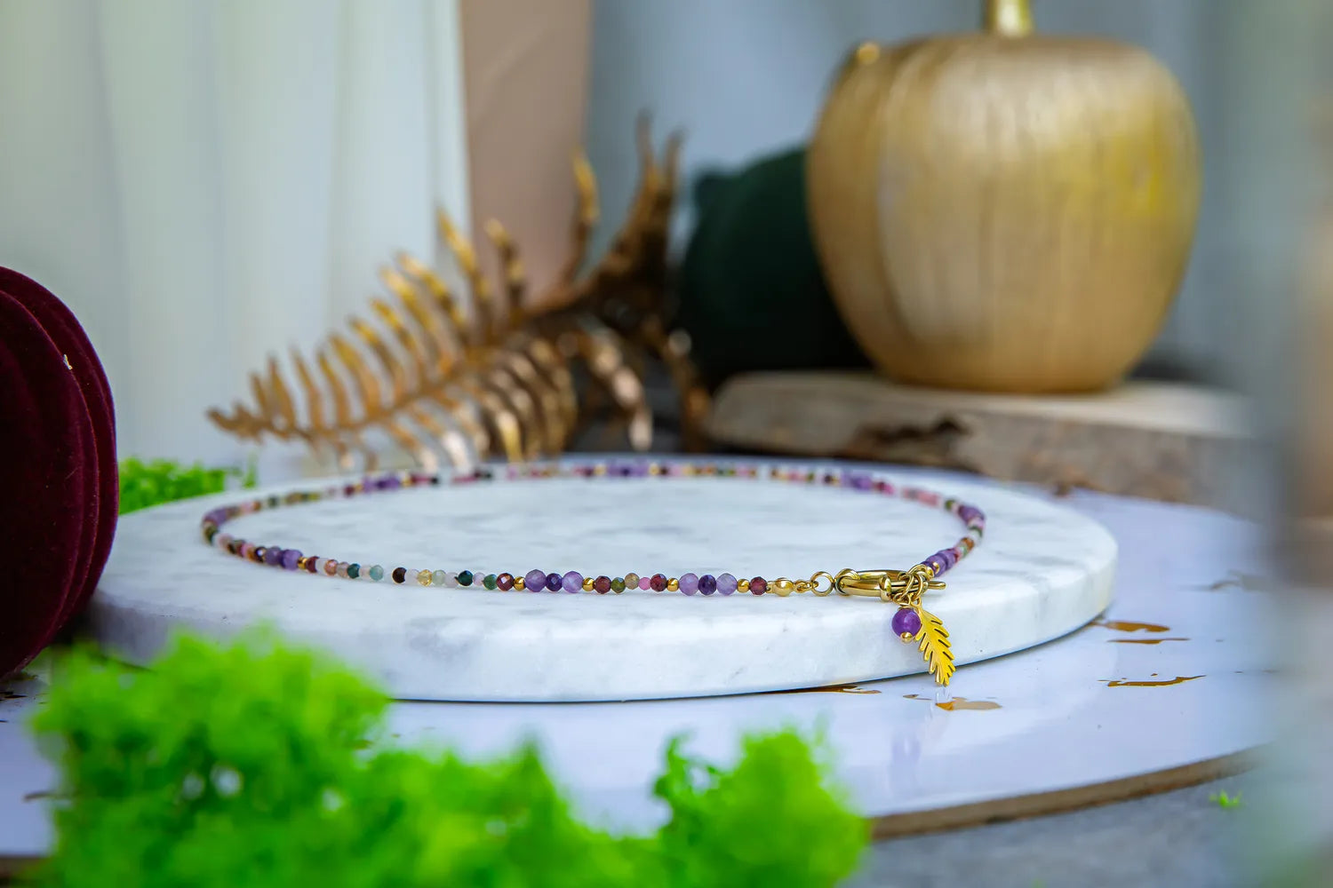 TĘCZOWA AURA - NASZYJNIK Z KOLOROWEGO TURMALINU I GRANATU, prezentowany na marmurowym talerzu z delikatnym złotym liściem zawieszką. W tle widoczne złote dekoracje i zielone liście.