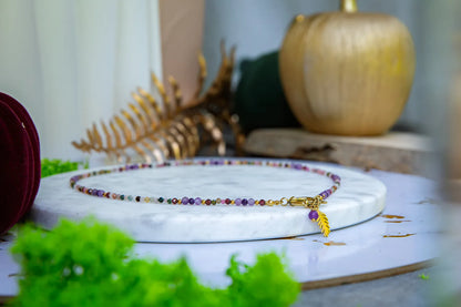 TĘCZOWA AURA - NASZYJNIK Z KOLOROWEGO TURMALINU I GRANATU, prezentowany na marmurowym talerzu z delikatnym złotym liściem zawieszką. W tle widoczne złote dekoracje i zielone liście.