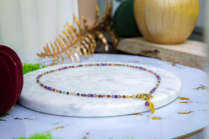 TĘCZOWA AURA - NASZYJNIK Z KOLOROWEGO TURMALINU I GRANATU, prezentowany na białym marmurowym podstawce. Naszyjnik składa się z kolorowych koralików turmalinu i granatu, z dodatkiem złotego zawieszka w kształcie liścia. W tle widoczne są złote liście, zielone liście, czerwona aksamitna torba i pomarańczowa dynia.