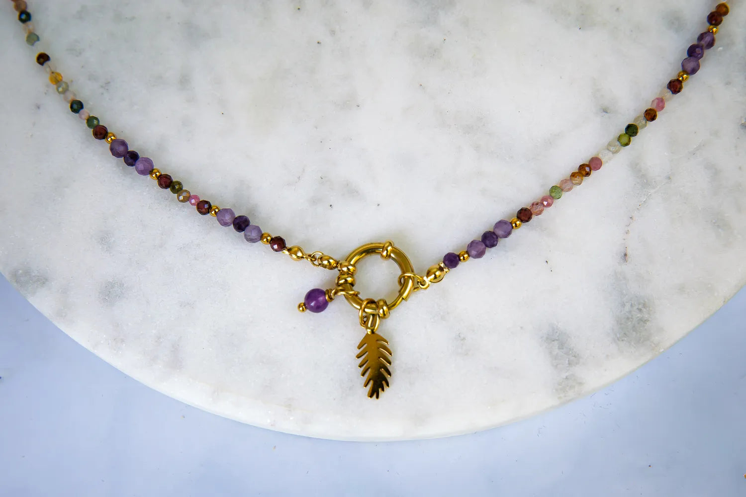 TĘCZOWA AURA - NASZYJNIK Z KOLOROWEGO TURMALINU I GRANATU. Naszyjnik z kolorowymi koralikami turmalinu i granatu, ozdobiony złotym pierścieniem i liściem, na białym marmurowym tle.