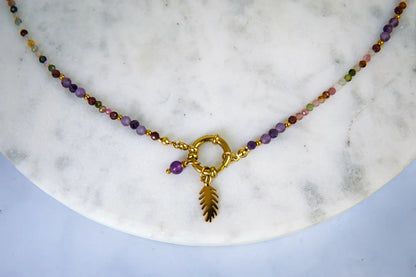 TĘCZOWA AURA - NASZYJNIK Z KOLOROWEGO TURMALINU I GRANATU. Naszyjnik z kolorowymi koralikami turmalinu i granatu, ozdobiony złotym pierścieniem i liściem, na białym marmurowym tle.