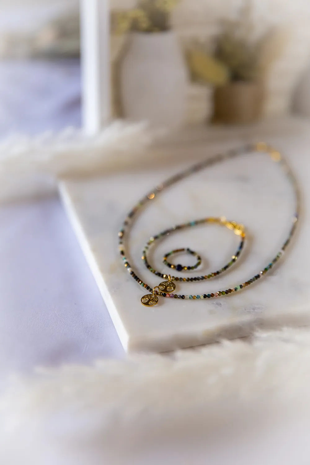 Tęczowa Harmonia Turmalin Drzewko Szczęścia: naszyjnik i pierścionek z kamieni turmalinowych na marmurowej podstawie, ozdobione złotymi zawieszkami Drzewka Szczęścia.