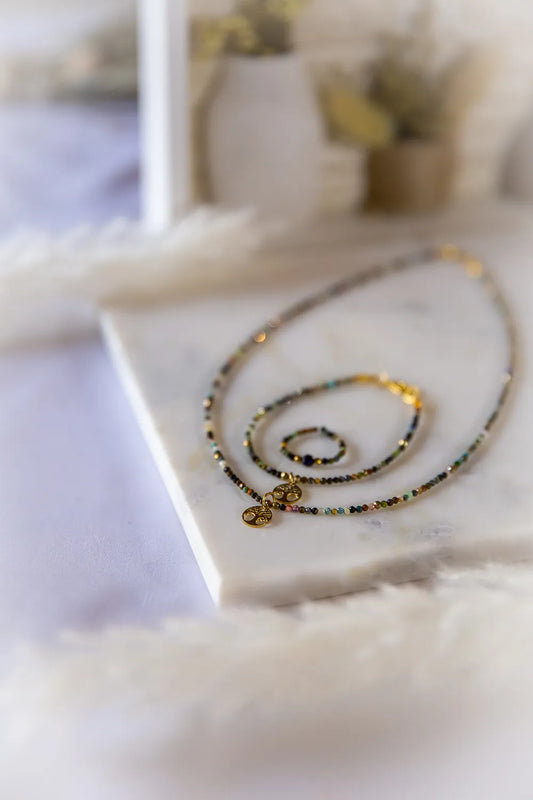 Tęczowa Harmonia Turmalin Drzewko Szczęścia: naszyjnik i pierścionek z kamieni turmalinowych na marmurowej podstawie, ozdobione złotymi zawieszkami Drzewka Szczęścia.