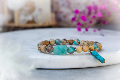 Turkusowa Oaza Bransoletka Z Jaspisu I Turkusu Koraliki: Bransoletka z koralików jaspisu, turkusu i złotymi przekładkami na marmurowym tle, z dominującymi odcieniami niebieskiego i beżu.