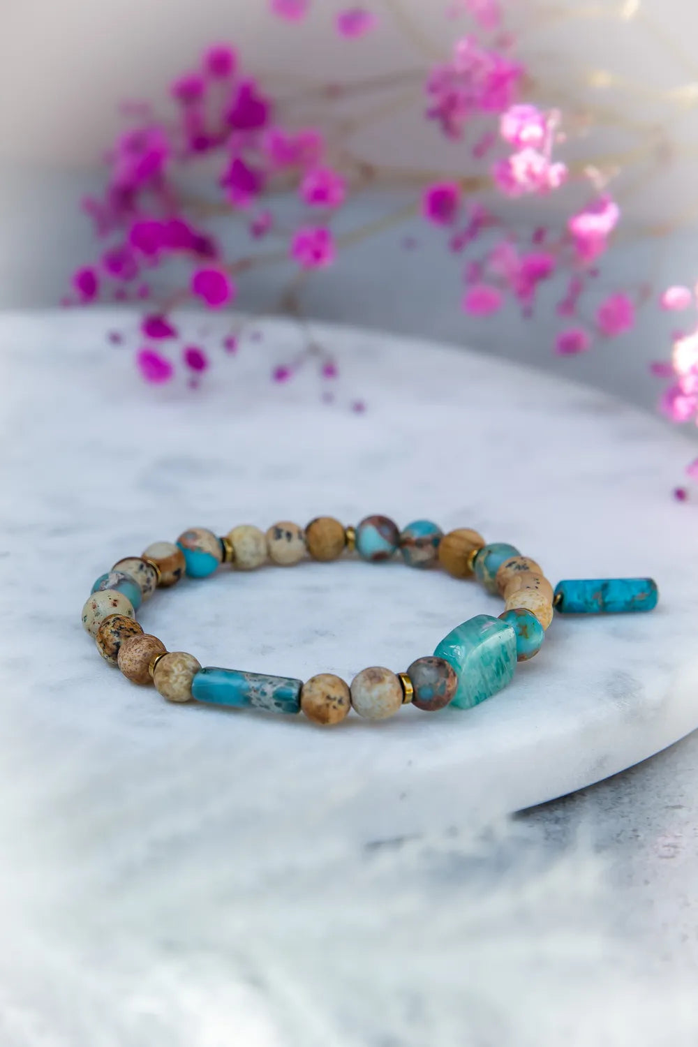 Turkusowa Oaza Bransoletka Z Jaspisu I Turkusu Kwiaty z tęczowym jaspisem i turkusowymi kamieniami na marmurowej powierzchni z różowymi kwiatami w tle.