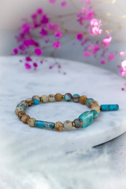 Turkusowa Oaza Bransoletka Z Jaspisu I Turkusu Kwiaty z tęczowym jaspisem i turkusowymi kamieniami na marmurowej powierzchni z różowymi kwiatami w tle.