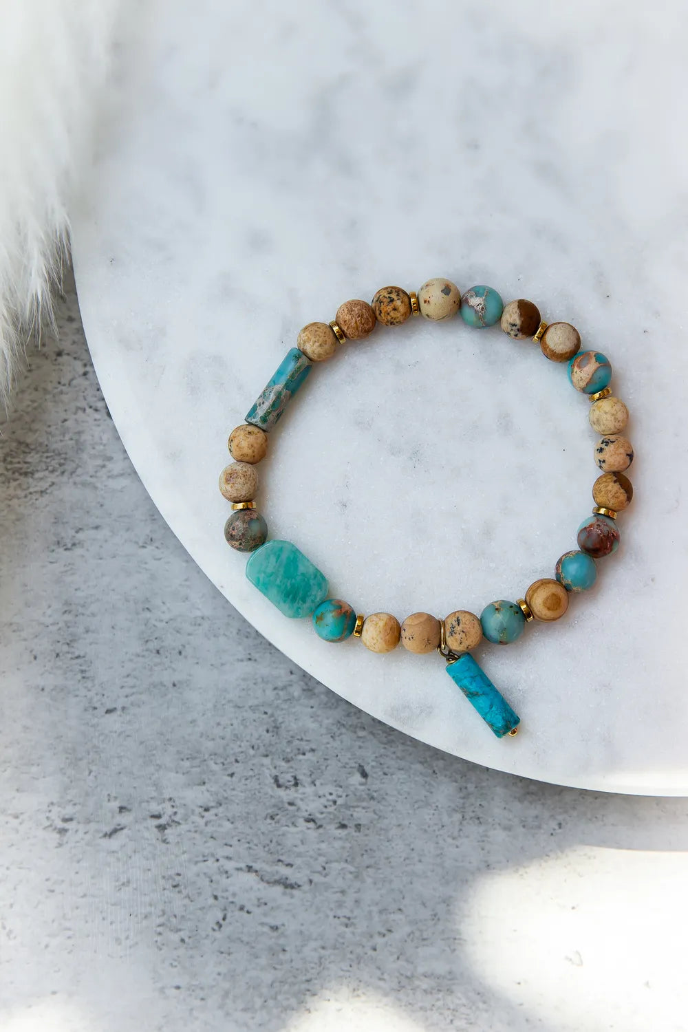 Turkusowa Oaza Bransoletka Z Jaspisu I Turkusu Marmur z płaskimi i okrągłymi kamieniami w odcieniach beżu, turkusu i brązu na białej powierzchni z przyciętym puszystym elementem po lewej.
