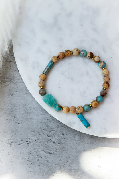 Turkusowa Oaza Bransoletka Z Jaspisu I Turkusu Marmur z płaskimi i okrągłymi kamieniami w odcieniach beżu, turkusu i brązu na białej powierzchni z przyciętym puszystym elementem po lewej.