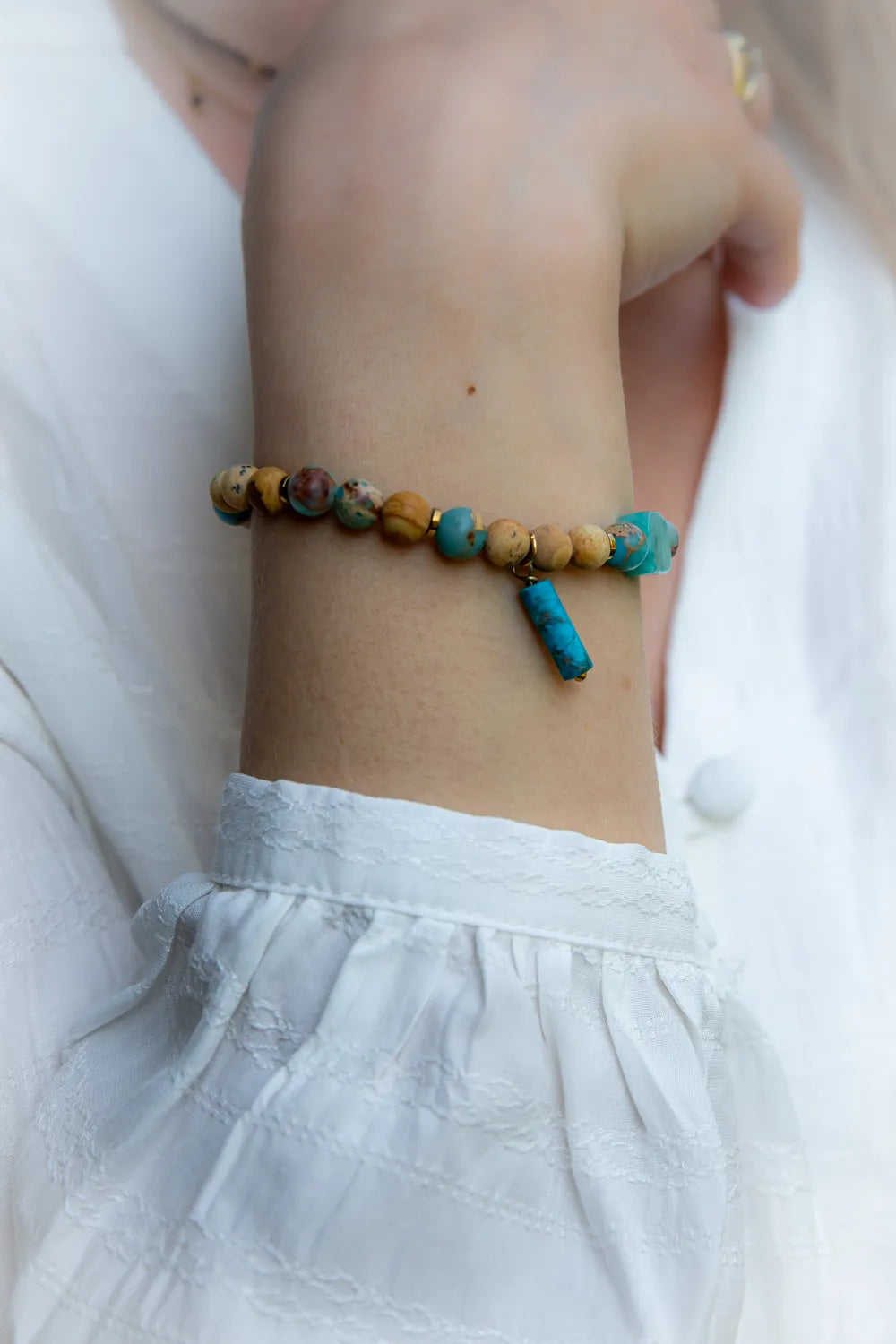 Turkusowa Oaza Bransoletka Z Jaspisu I Turkusu Ozdobna: Bransoletka z koralików z turkusu, jaspisu i złotych przekładek, z wiszącym, turkusowym walcem. Zdjęcie na tle białej, lekko rozmazanej tkaniny.